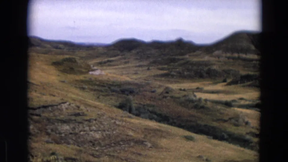 1967 Yellowstone landscape frame with sloped dry grass, rocky ground, brush, and darker distant landforms.