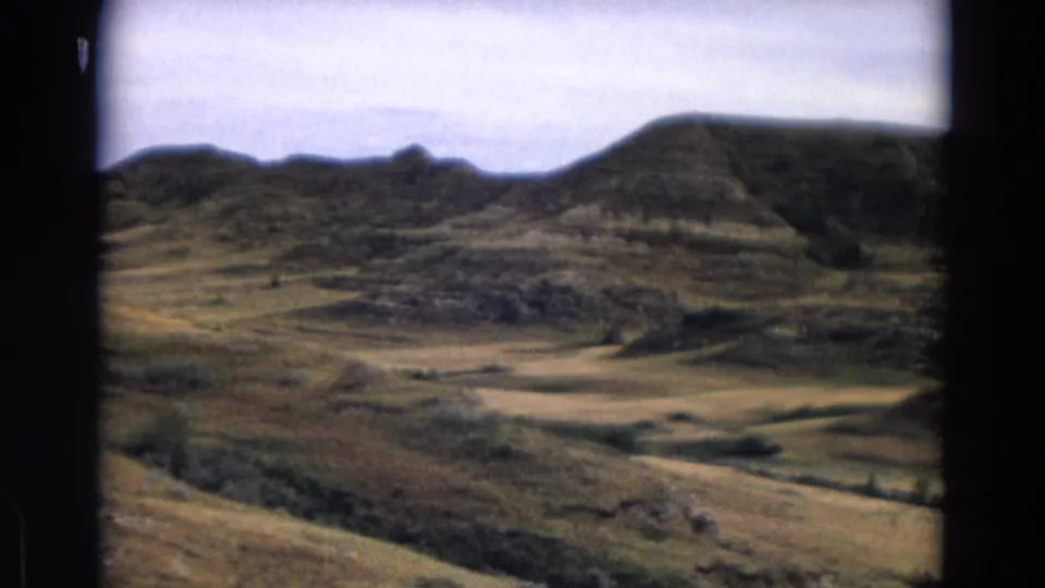 1967 Yellowstone home-movie frame showing a wider panorama of rolling terrain, eroded rock bands, shrubs, and a rounded hilltop.