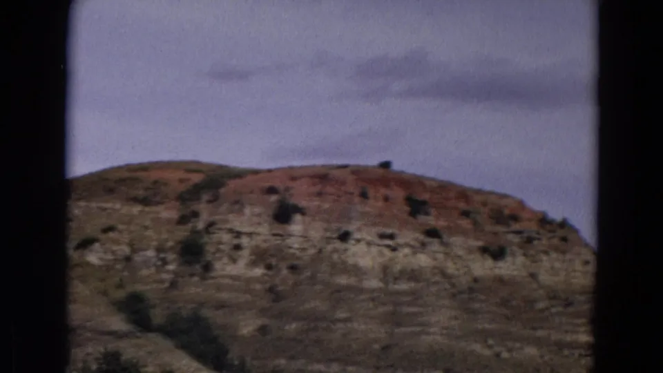 1967 Yellowstone home-movie frame showing a dry layered rock slope with sparse shrubs beneath a pale blue sky.