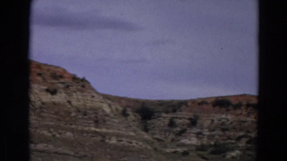 1967 Yellowstone home-movie view of dry layered terrain, low ridges, and sparse vegetation under hazy blue sky.