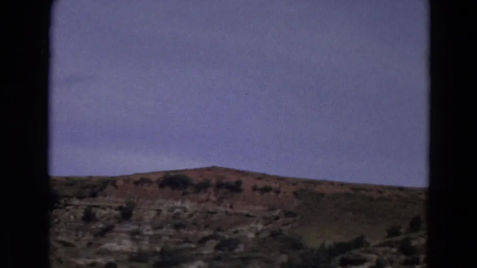 1967 Yellowstone frame showing a distant rocky ridge, dry foreground slope, scattered shrubs, and broad pale sky.