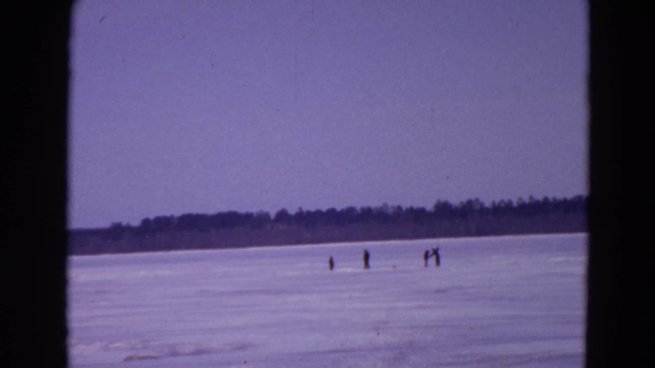 Frozen lake with small distant figures on the ice and a dark treeline under a pale sky.