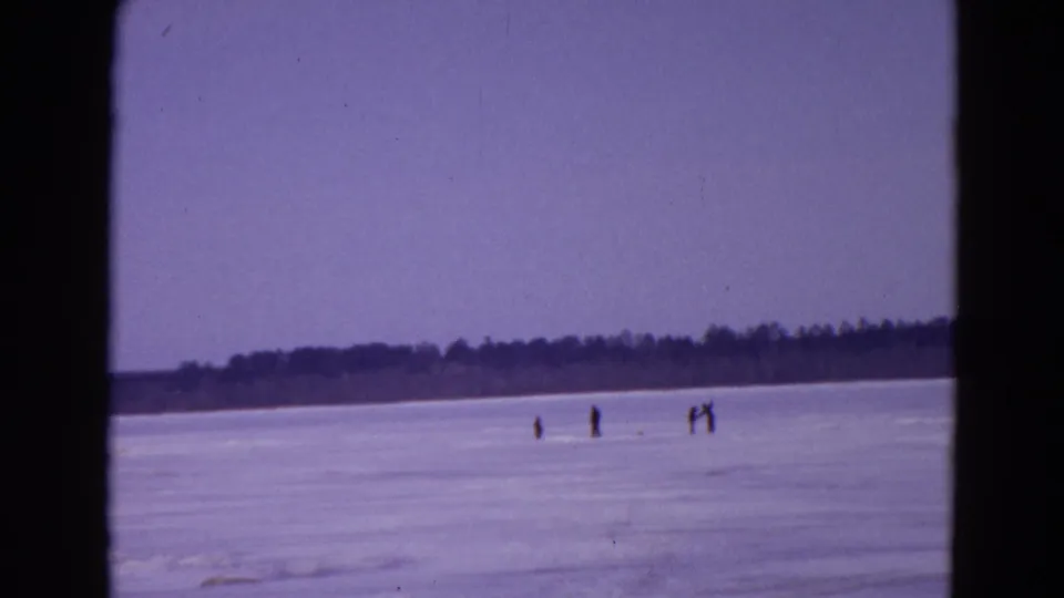 1967 Yellowstone home-movie frame showing distant people on a frozen lake beneath a pale sky and dark treeline.