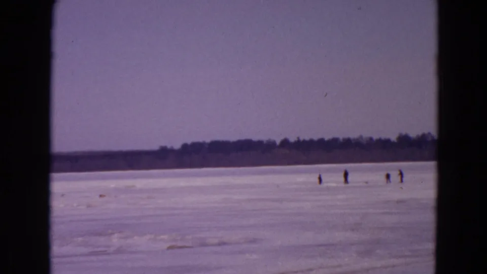 1967 8mm home-movie frame showing a frozen lake in Yellowstone Park, Wyoming, with distant figures on the ice and a low wooded shoreline.