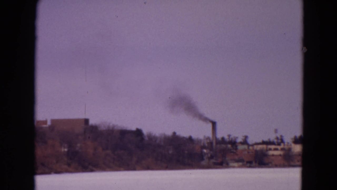 1967 8mm home-movie frame of a frozen lake with distant industrial buildings and a smokestack emitting dark smoke near the shoreline.