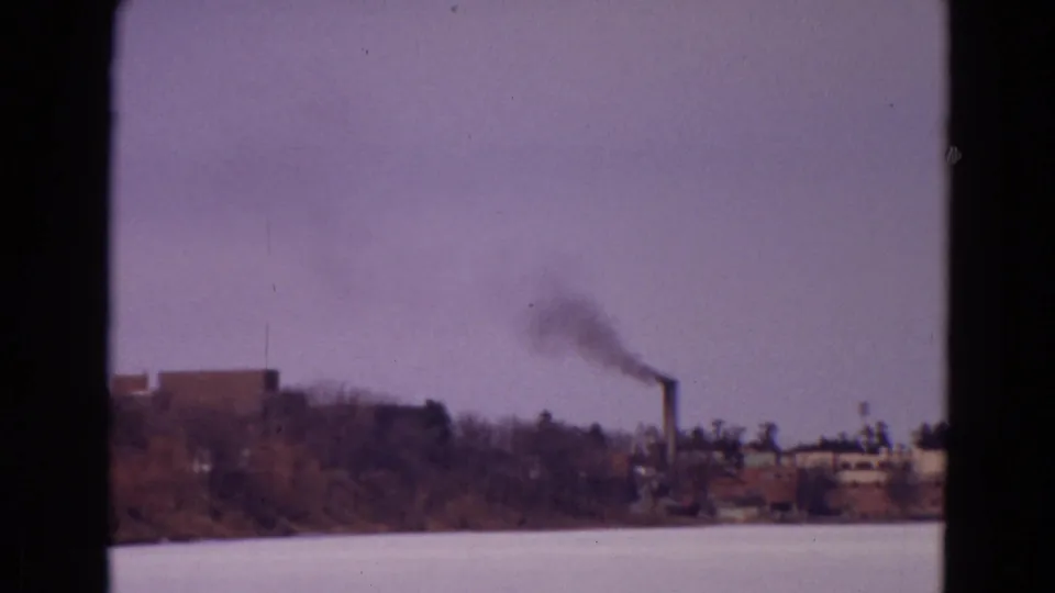 1967 8mm home-movie frame of a frozen lake with distant industrial buildings and a smokestack emitting dark smoke near the shoreline.