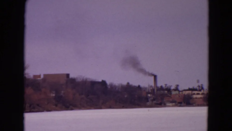 1967 8mm home-movie frame showing the frozen lake foreground and a distant smokestack with dark smoke above low industrial structures.