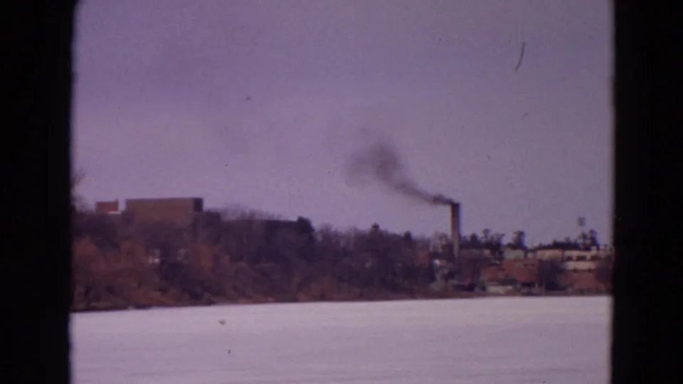 1967 8mm home-movie frame of a dark smoke plume rising from a distant stack beyond a frozen lake in Yellowstone Park, Wyoming.