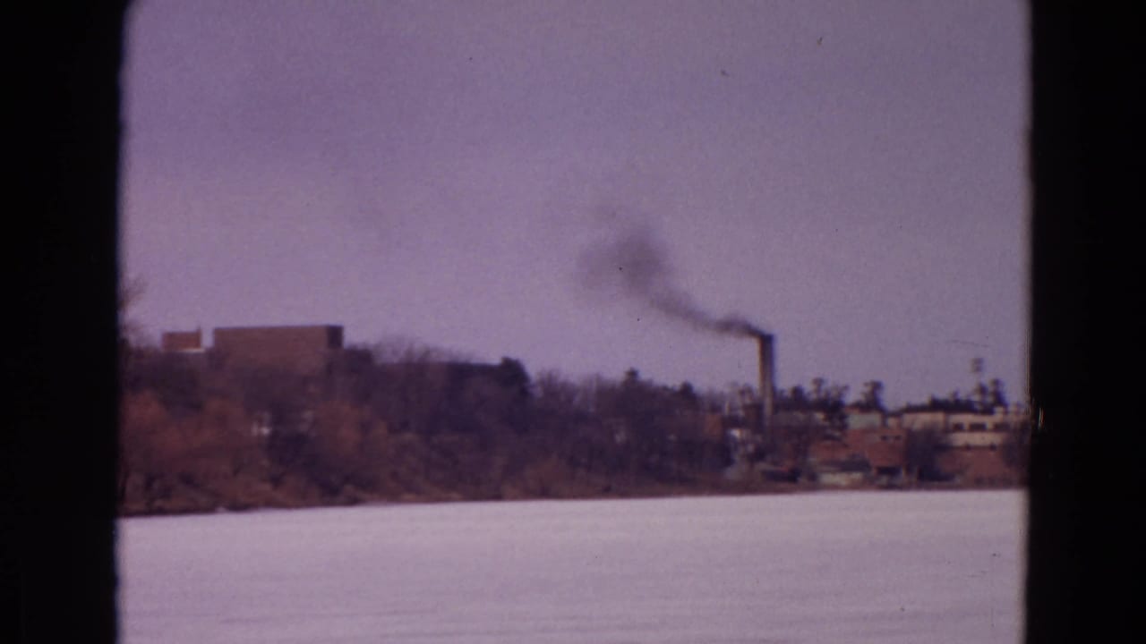 Distant smokestack releasing dark smoke beyond a frozen lake.