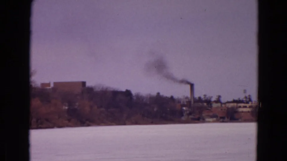 1967 Wyoming home-movie frame with a smokestack and dark smoke seen across a frozen lake through dark borders.