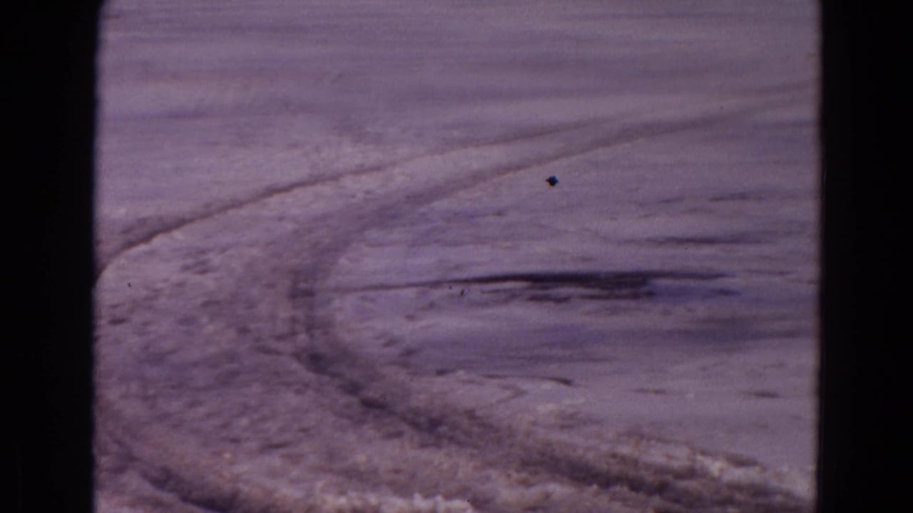 Curving tire tracks cross a broad snow-covered open area with a distant dark tree line.