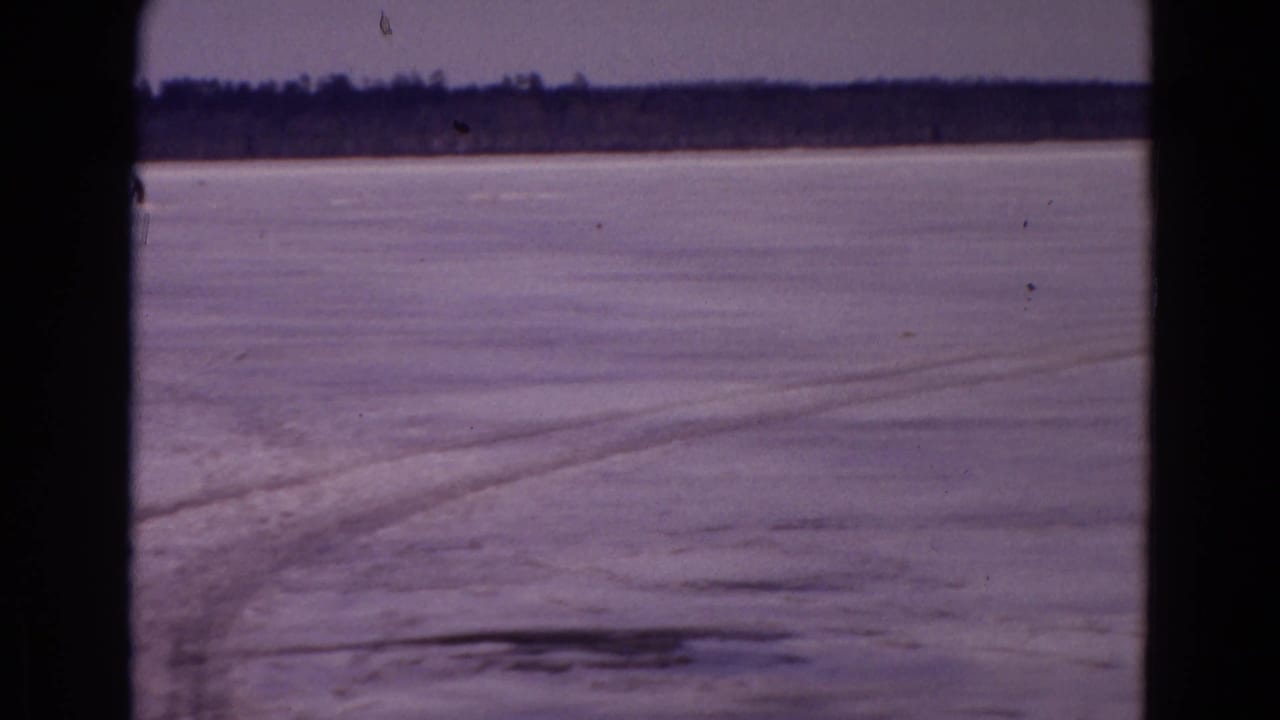 The camera looks across a frozen Yellowstone snowfield with faint track lines leading toward a distant tree line.