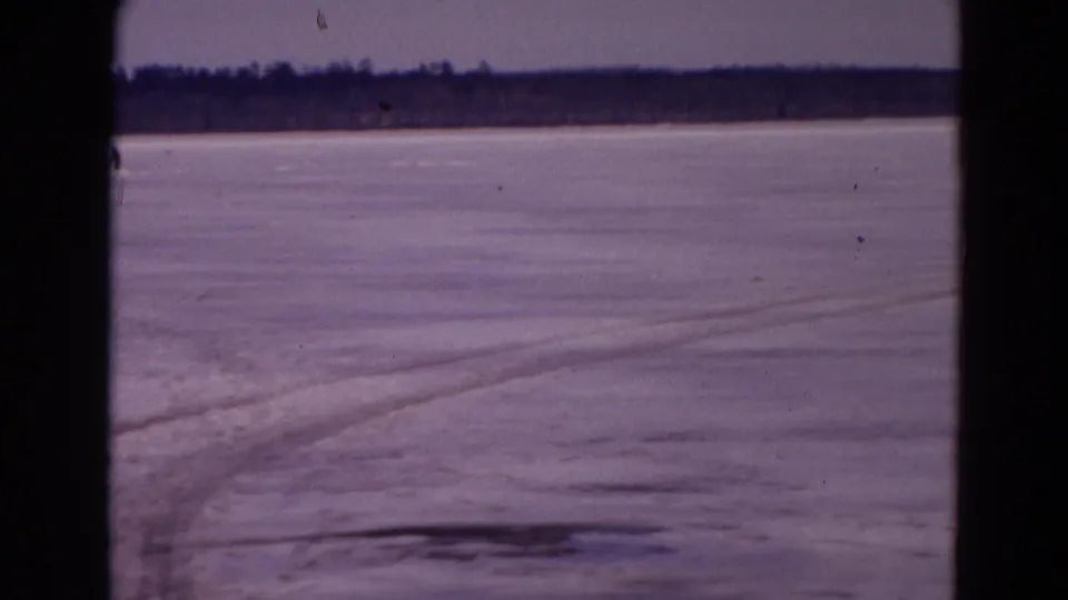 The camera looks across a frozen Yellowstone snowfield with faint track lines leading toward a distant tree line.