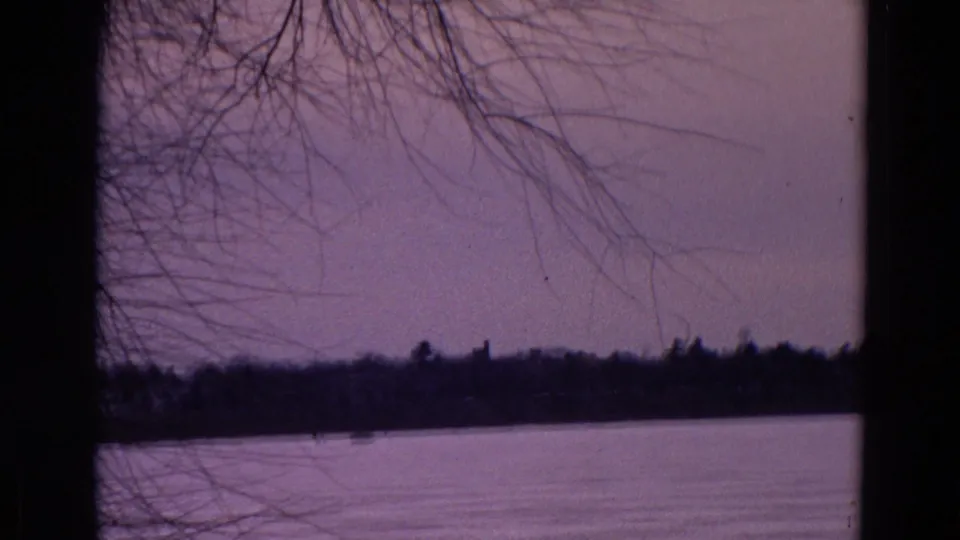 1967 Yellowstone frame with leafless branches above a calm lake or river and a dark shoreline under a purple evening sky.