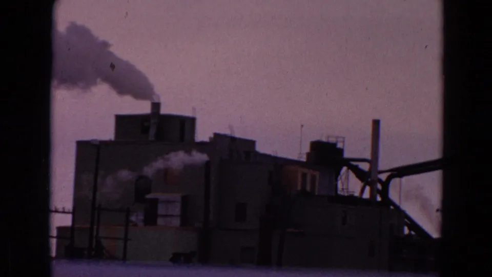 1967 8mm footage shifts to an industrial complex with boxy buildings, smokestacks, and smoke plumes rising into a pale sky.