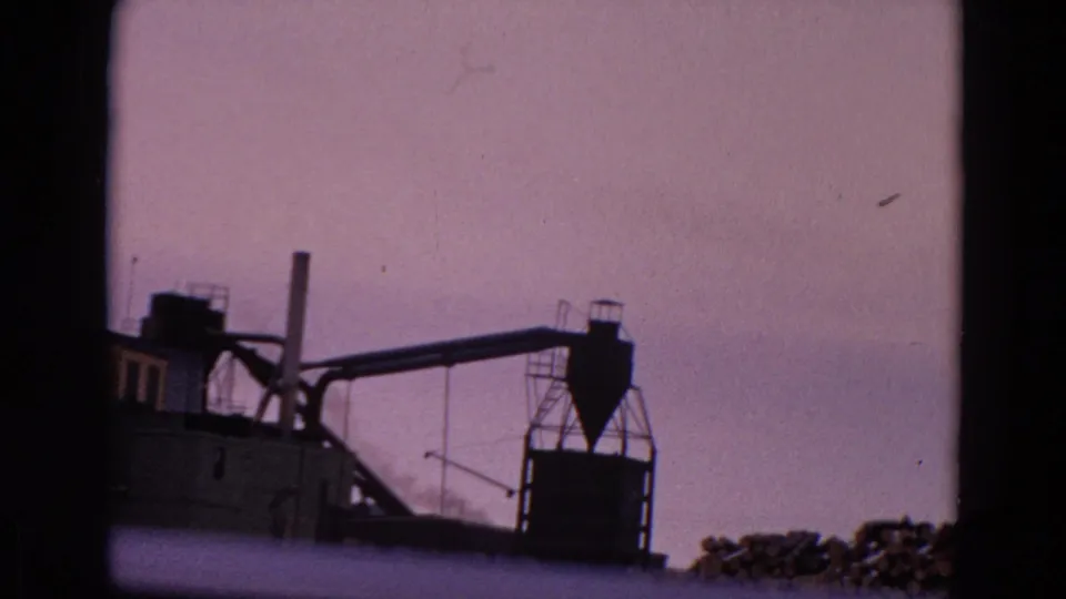 A vintage 8mm view of an industrial plant shows conveyor-like structures, a tower, a smokestack, and low smoke near the buildings.