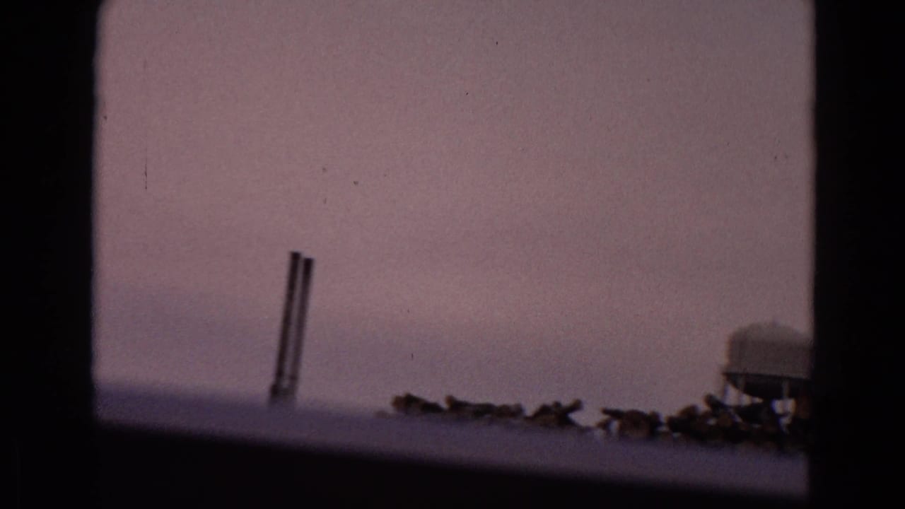 Window-framed view of distant pipes, low structures, timber piles, and a small tank.