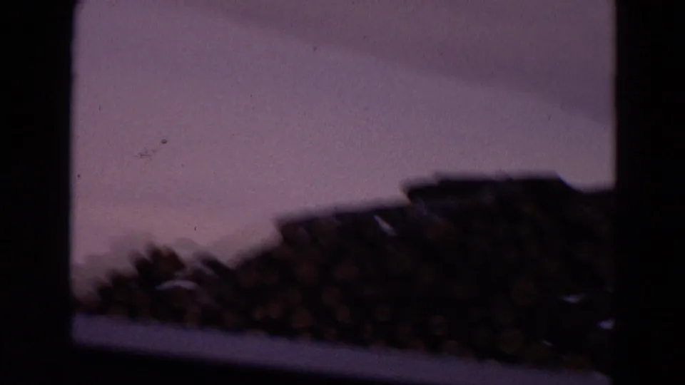 A soft, low-light 1967 8mm frame holds on timber piles and distant industrial ground beyond the window edge.
