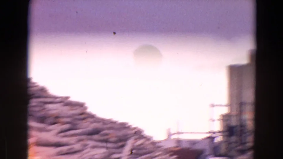 1967 8mm frame showing a lumberyard view with stacked logs, a dark fence or post, and a small structure in bright exposure.