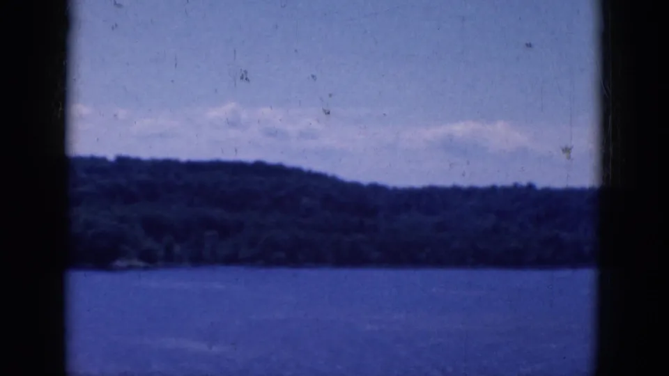 1968 8mm home-movie frame showing open water, a forested hill, pale sky, and dark window bars in the foreground.