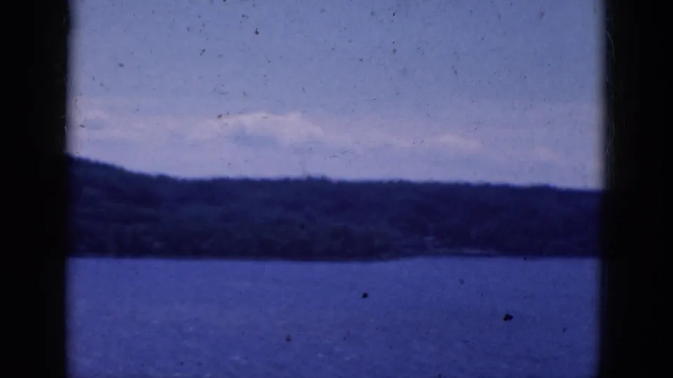 Blue 8mm frame of a broad water surface and tree-covered shoreline, partially blocked by dark vertical window edges.