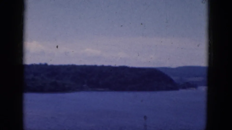 1968 home-movie frame with blue water, a dark wooded point, distant hills, and window-like vertical framing.