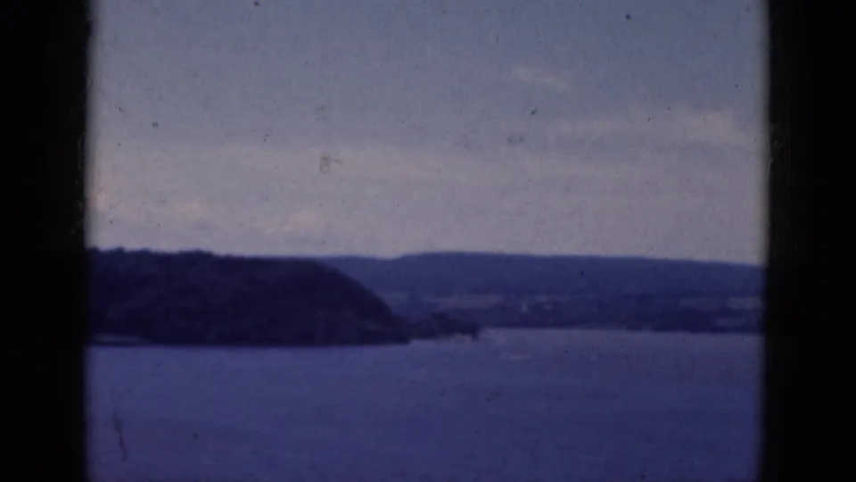 Blue-tinted 8mm frame showing open water, a forested shoreline, distant hills, pale clouds, and dark frame edges.