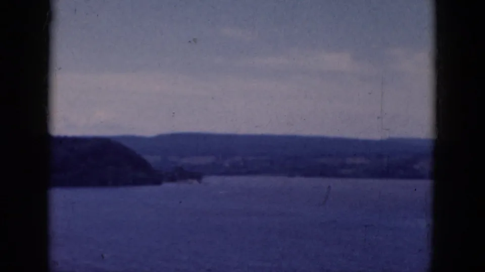 1968 8mm frame showing blue lake water, far shoreline, and low hills viewed through dark window bars.