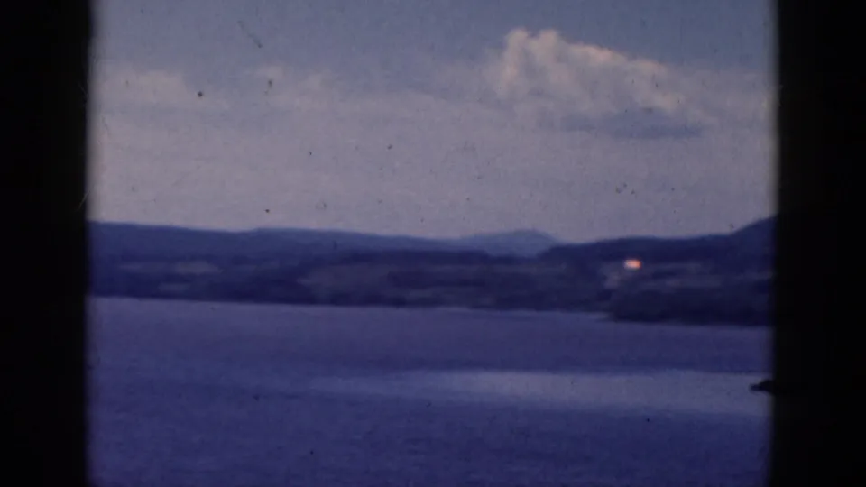 1968 8mm frame with lake water, layered hills, soft clouds, and a small distant light near the shoreline.