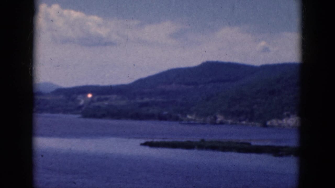 Calm lake below wooded hills with a low island or peninsula in the foreground.