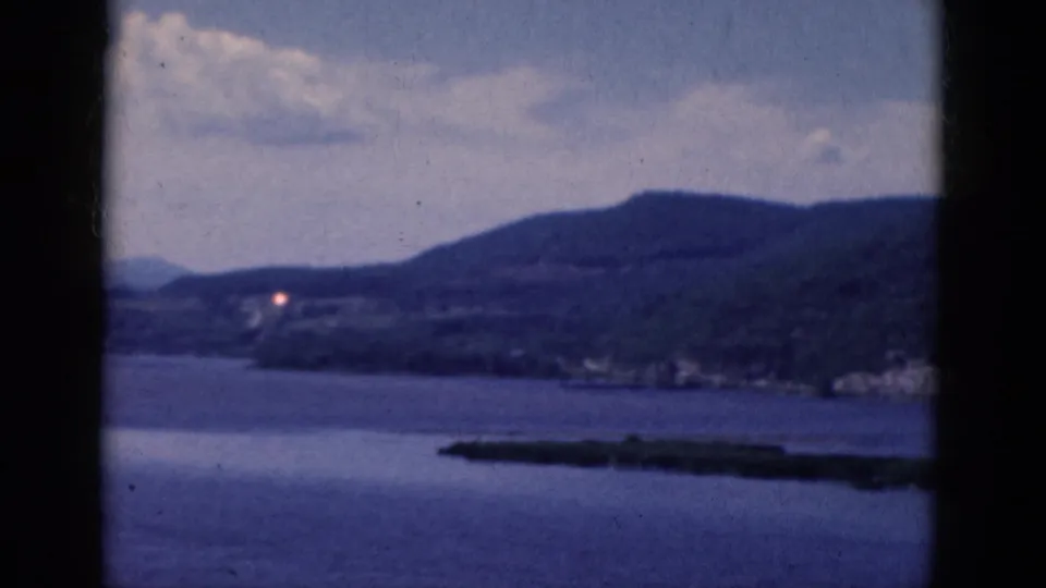 1968 8mm home-movie frame showing blue lake water, wooded hills, distant shoreline, and a low island or peninsula under a partly cloudy sky.