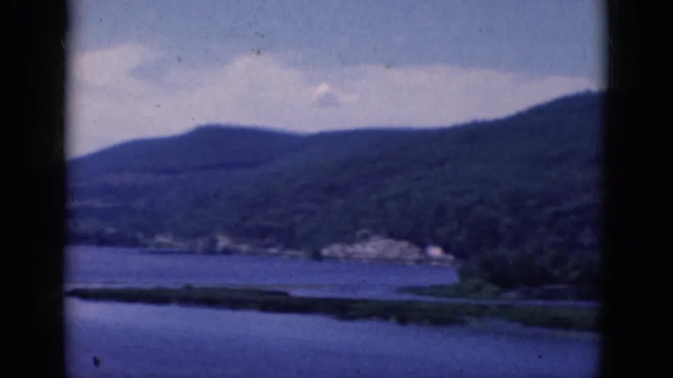 1968 8mm frame of a broad lake and forested hills, with distant shoreline structures and a dark vertical gate or scan divider through the view.