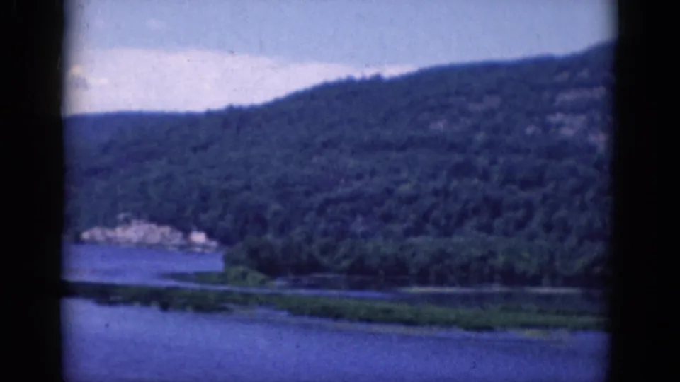 1968 8mm home-movie frame showing a wooded hillside, blue lake water, low marshy shoreline, and a small cluster of distant light-toned structures.