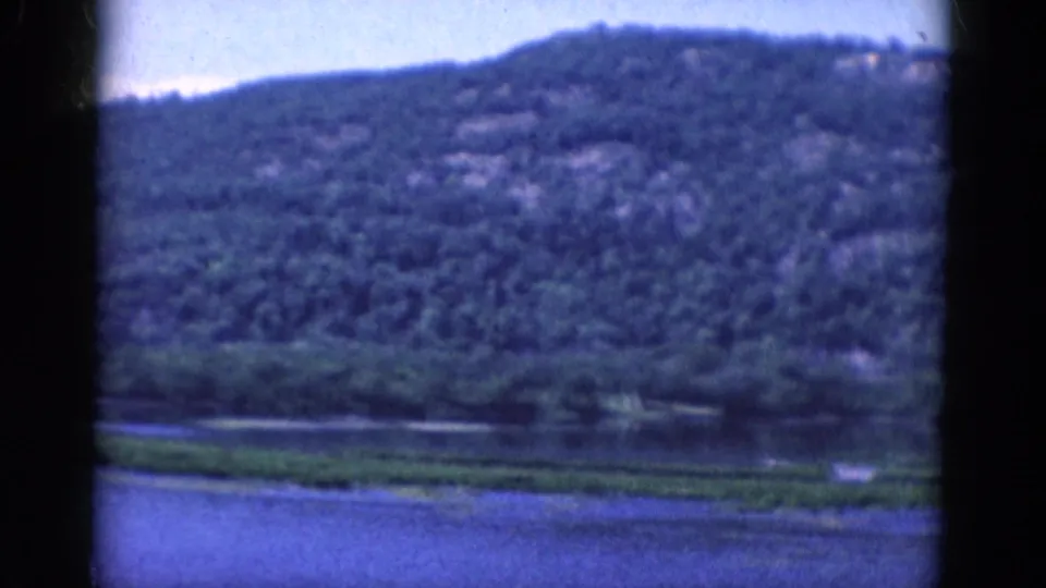 1968 8mm frame of a wooded slope and lake edge, with a low strip of shoreline vegetation and visible film grain and gate shadow.