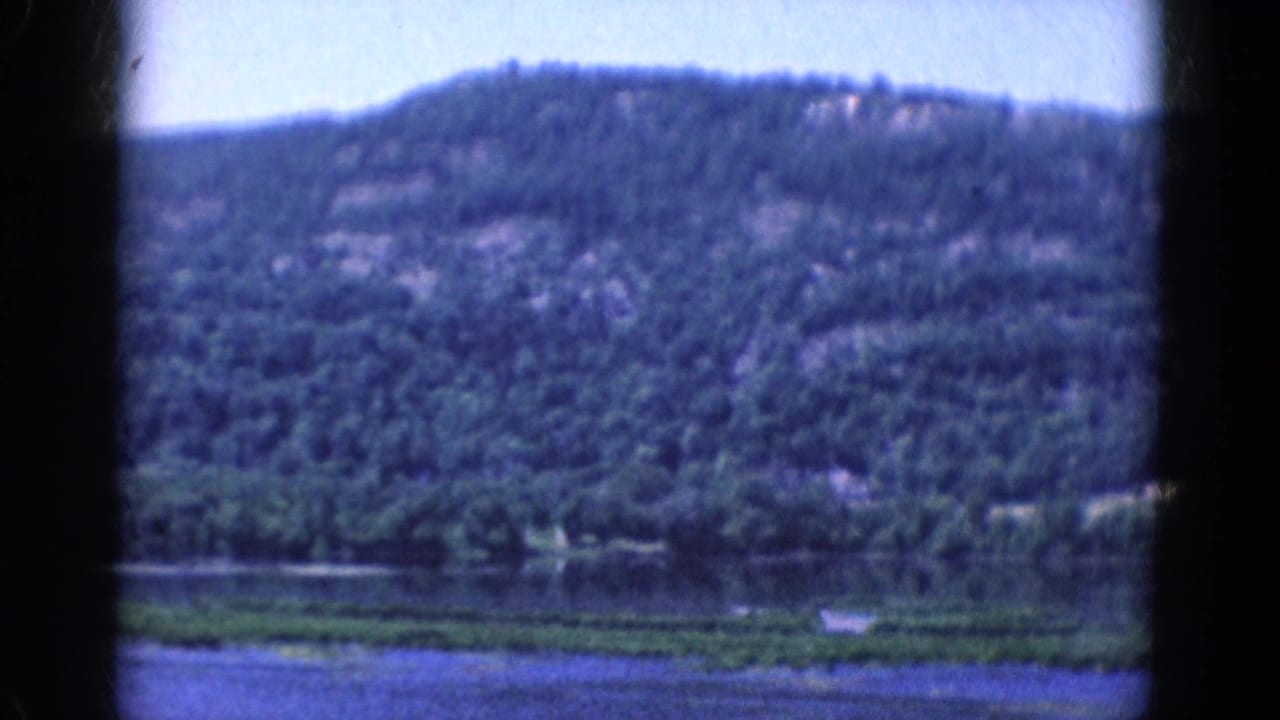 1968 8mm home-movie frame showing water, shoreline vegetation, and a wooded hill under pale blue sky.