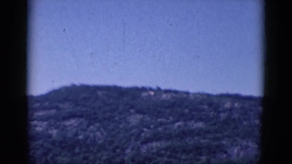 Soft 1968 8mm landscape frame showing the upper slope of a wooded hill against blue sky.