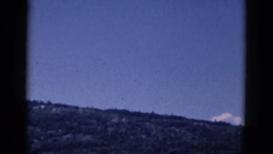 1968 8mm frame showing a distant wooded hillside beneath a blue sky with dark gate edges and soft film grain.