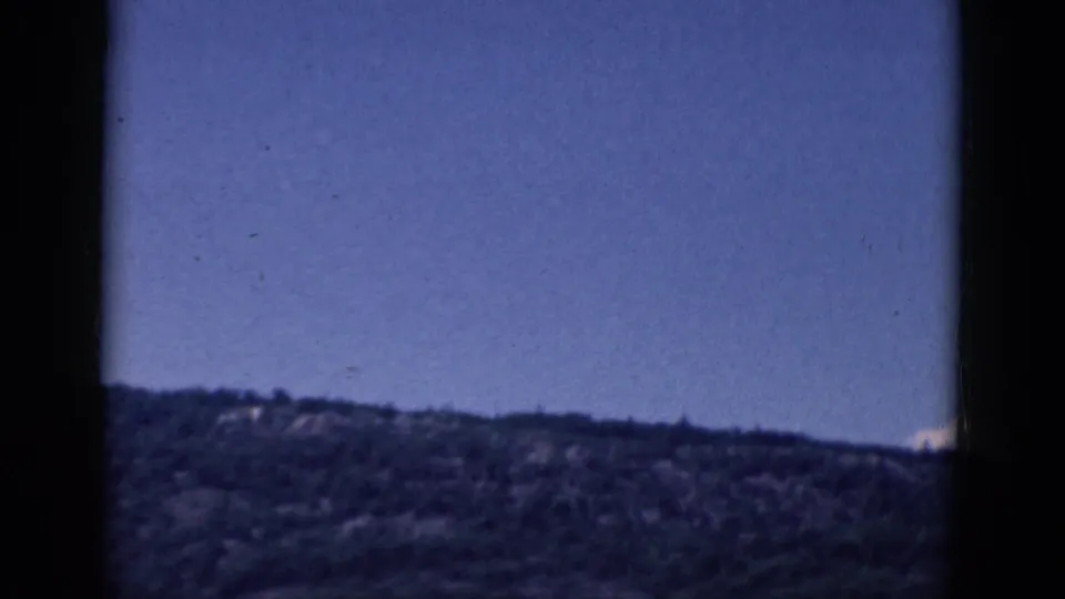 1968 8mm frame of a tree-covered ridge beneath open blue sky, bordered by dark film-gate edges.