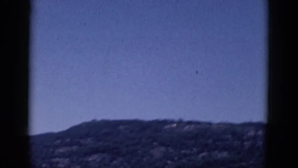 1968 8mm frame with a low wooded hillside, broad blue sky, soft focus, and dark frame edges.