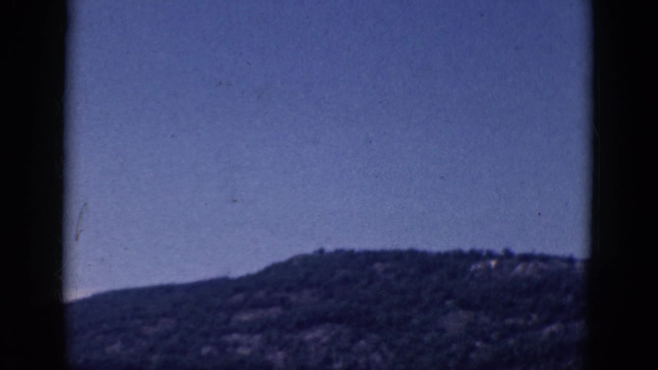 Dark wooded hillside under a blue sky, framed by black film edges.