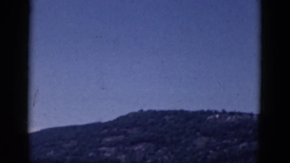 1968 8mm frame showing a distant wooded hillside under blue sky, with heavy dark film-gate borders and no people visible.