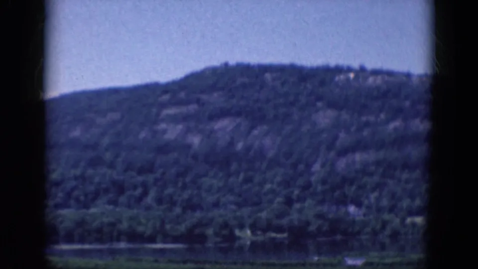 A distant wooded hillside rises above a calm lake edge in this 1968 8mm home-movie frame, with no people or signage visible.