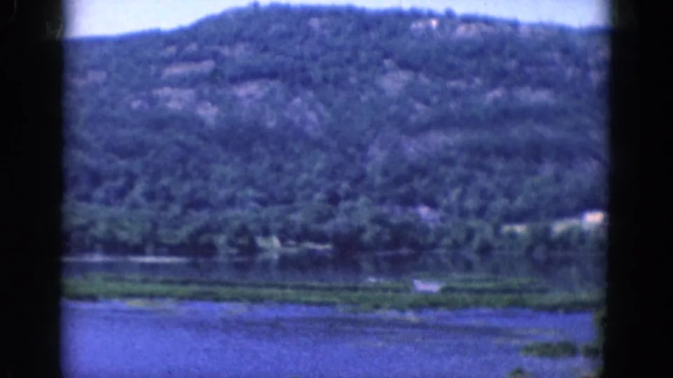 A green treed hillside overlooks lake water and low shoreline vegetation in a softly focused 1968 8mm landscape frame.