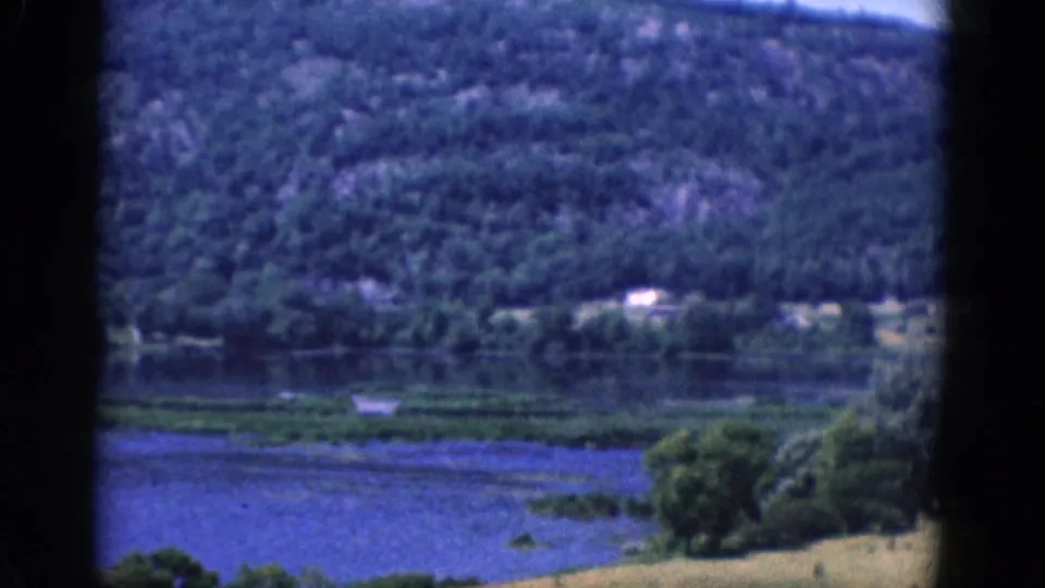 The final frame shows calm lake water, a grassy shoreline, and a wooded hillside with small distant light structures, captured on 1968 8mm film.