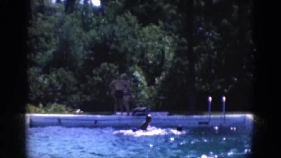 Swimmers move through an outdoor pool while a person stands on the deck near dense trees and pool ladders.