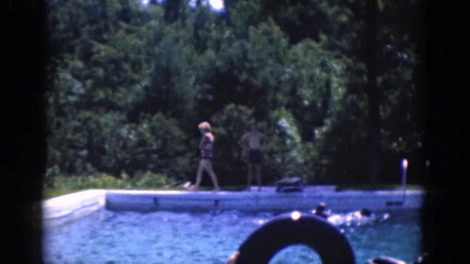 A poolside figure walks past a life preserver as swimmers remain in the water below a wooded background.