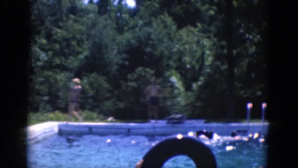 Swimmers cluster near the pool edge while a large life preserver and wooded background frame the home-movie view.
