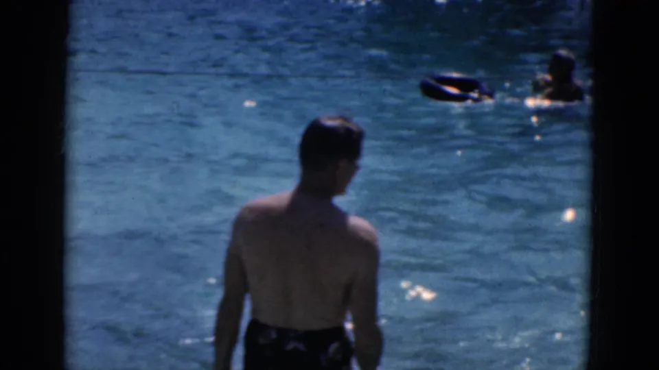 1968 8mm home-movie frame of a man by a swimming pool while other people float and swim in sunlit water.