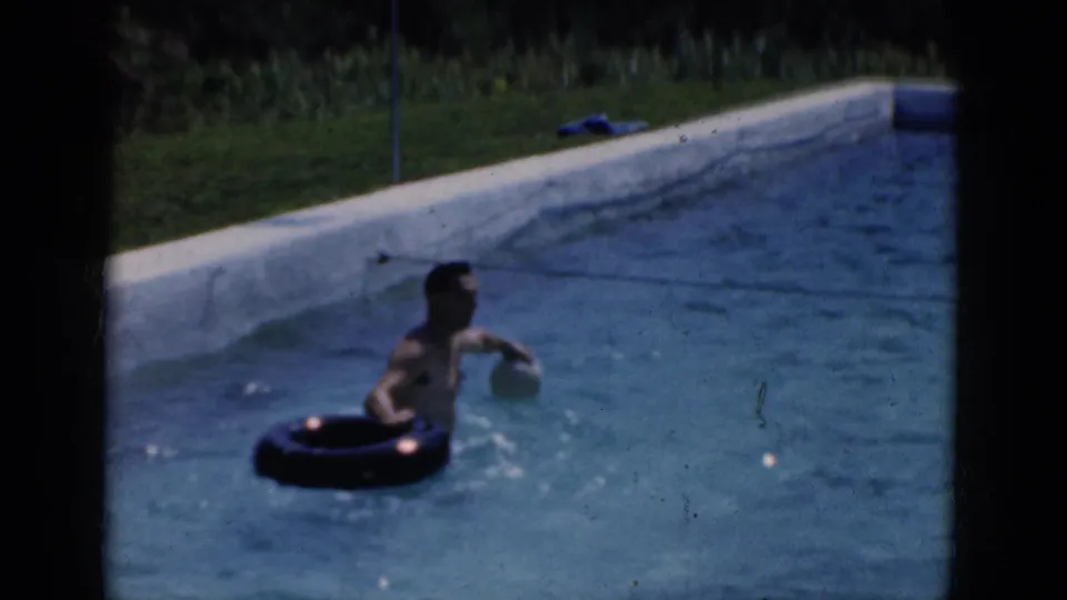 Home-movie frame of a man in a swimming pool with a dark inner tube, pale pool wall, grass, and sunlit water.