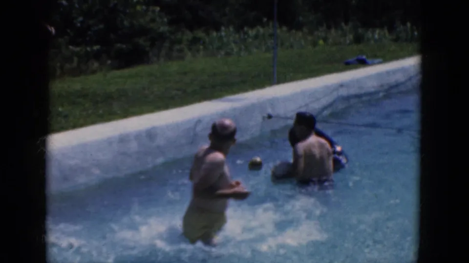 1968 8mm home-movie frame of two people splashing and playing in a pool near a ball and grassy edge.